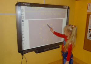 Dziewczynka stoi pod tablicą interaktywną i przelicza w fladze Unii Europejskiej gwiazdki.